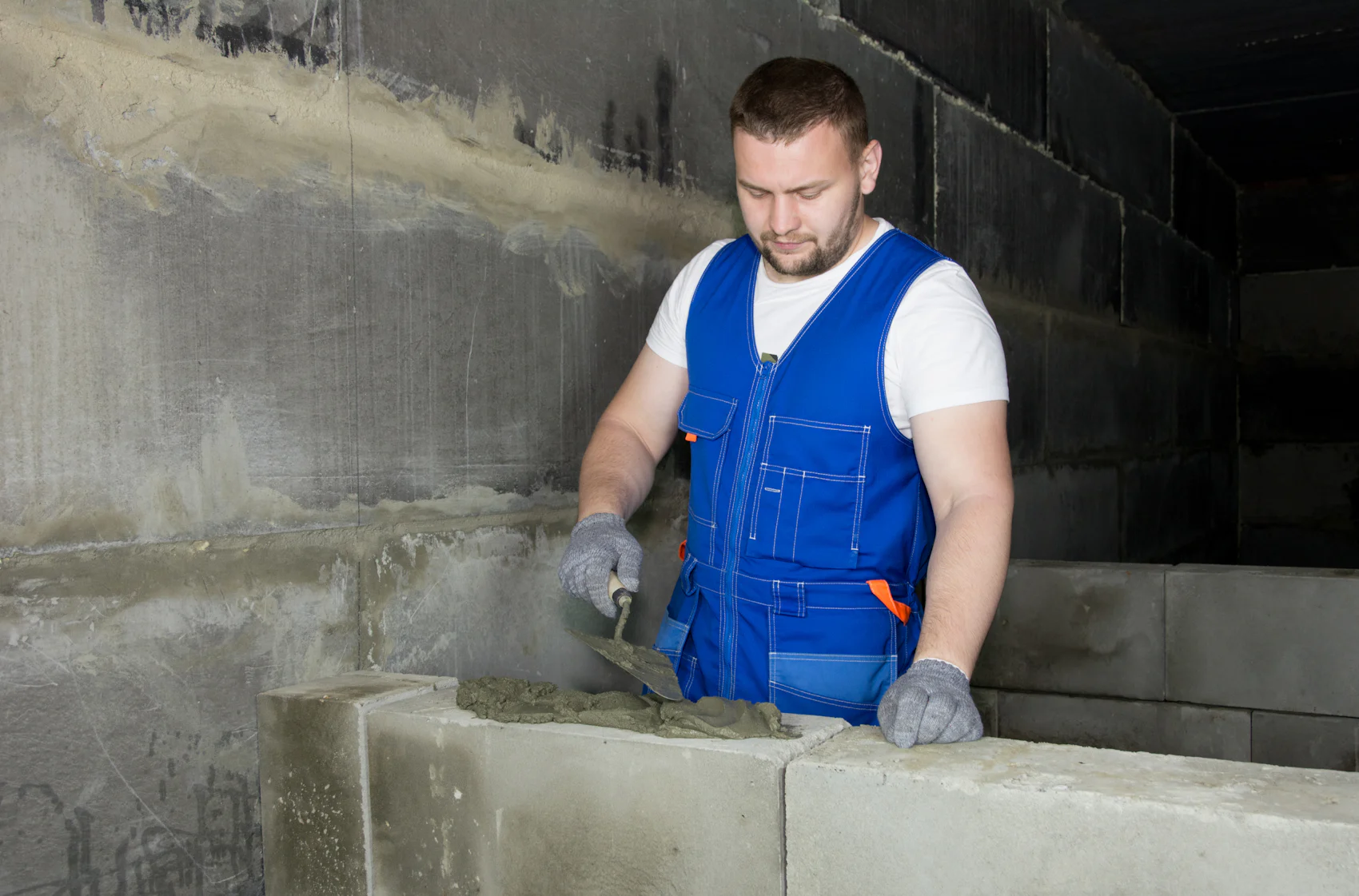 Ein Handwerker im blauen Latz-Overall zieht Mörtel mit der Kelle über eine Wand aus Betonsteinen.